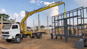 caminhão VW branco com guindaste Rodomunk GRM 16000 levantando módulo de estrutura metálica cinza com cinta amarela; trabalhadores guiam o alinhamento da peça em canteiro com vários pilares de concreto ao fundo.