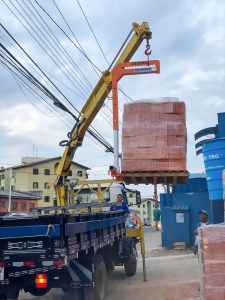 guindaste veicular (Munck) Rodomunk içando um pallet grande de matéria-prima (blocos/tijolos cerâmicos envoltos em filme) usando acessório tipo garfo/levantador de carga laranja com indicação de “2000 kg”; operação em rua com muitos cabos aéreos e trabalhadores acompanhando o descarregamento na carroceria do caminhão.