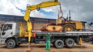 munck Rodomunk amarelo carregando máquina/equipamento pesado sobre caminhão prancha, preso por cintas; trabalhador de uniforme verde orienta a manobra ao lado da carga, com estabilizadores apoiados no chão e galpão industrial ao fundo.