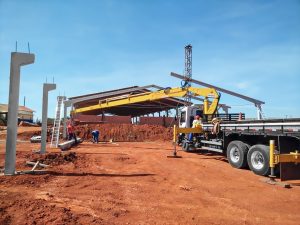 guindaste veicular Rodomunk com lança longa operando em obra de solo vermelho, posicionando/transportando perfis e elementos da estrutura enquanto equipe trabalha no canteiro; ao fundo, estrutura de cobertura em montagem e torre metálica vertical, com caminhão estabilizado por sapatas.