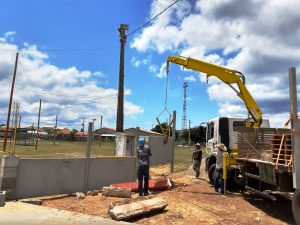 caminhão com munck Rodomunk amarelo içando e posicionando peça pré-moldada de concreto (painel/elemento de muro) com acessório de pega, em área aberta com postes e fiação; dois trabalhadores em solo orientam o encaixe ao lado de mureta e base de concreto.