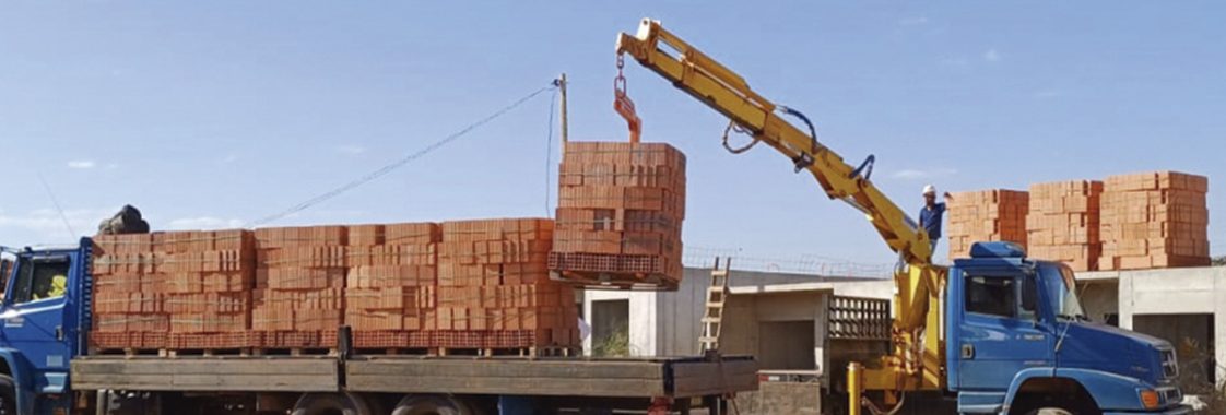 Guindaste veicular levantando carga de tijolos em obra, operação de içamento que exige comunicação do sinaleiro para orientar o operador.