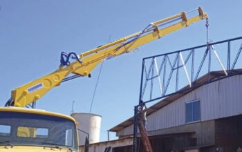 Guindaste veicular com lança estendida instalando estrutura metálica no topo de um galpão, demonstrando o alcance horizontal do equipamento sem reposicionar o caminhão.