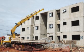 Guindaste veicular realizando o carregamento de peças pré-moldadas em um canteiro de obras, com patolas estabilizadas e operação próxima a um edifício em construção, ilustrando a etapa de içamento e posicionamento seguro das estruturas.