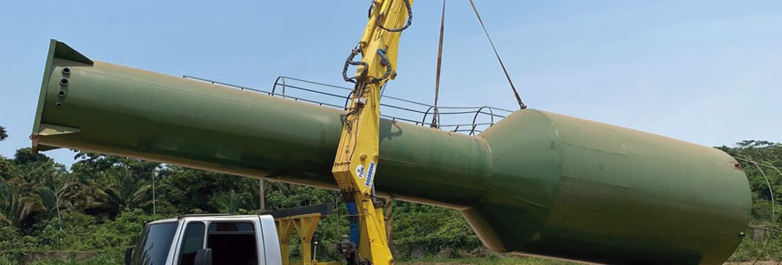 Guindaste veicular acoplado a caminhão realizando o içamento de uma caixa d’água de grande porte para posicionamento preciso na obra, demonstrando o uso do equipamento para elevação segura e controlada do reservatório.