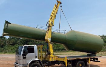 Guindaste veicular acoplado a caminhão realizando o içamento de uma caixa d’água de grande porte para posicionamento preciso na obra, demonstrando o uso do equipamento para elevação segura e controlada do reservatório.