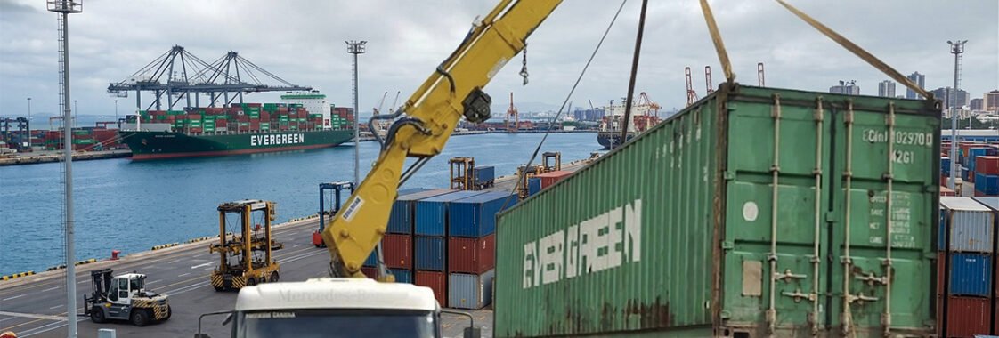 Guindaste veicular montado sobre caminhão movimentando um contêiner no pátio de um porto, com navio cargueiro e pilhas de contêineres ao fundo, ilustrando a mobilidade e o suporte logístico desse tipo de equipamento nas operações portuárias.