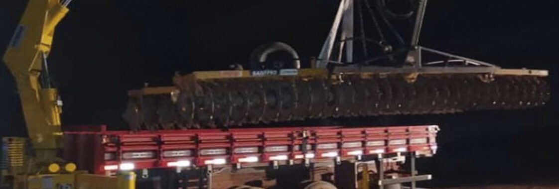 Guindaste veicular realizando içamento noturno de estrutura metálica sobre caminhão, com uso de iluminação artificial para garantir visibilidade e segurança na operação.