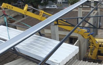 Guindaste veicular operando em espaço urbano reduzido, içando chapas metálicas sobre estrutura próxima a telhados e edificações, demonstrando a necessidade de precisão, controle de alcance e atenção a obstáculos ao redor.